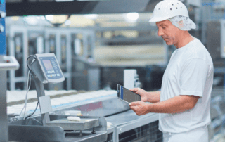 Factory worker standing by a bench scale with a mobile tablet, analysing Overall Equipment Effectiveness data.